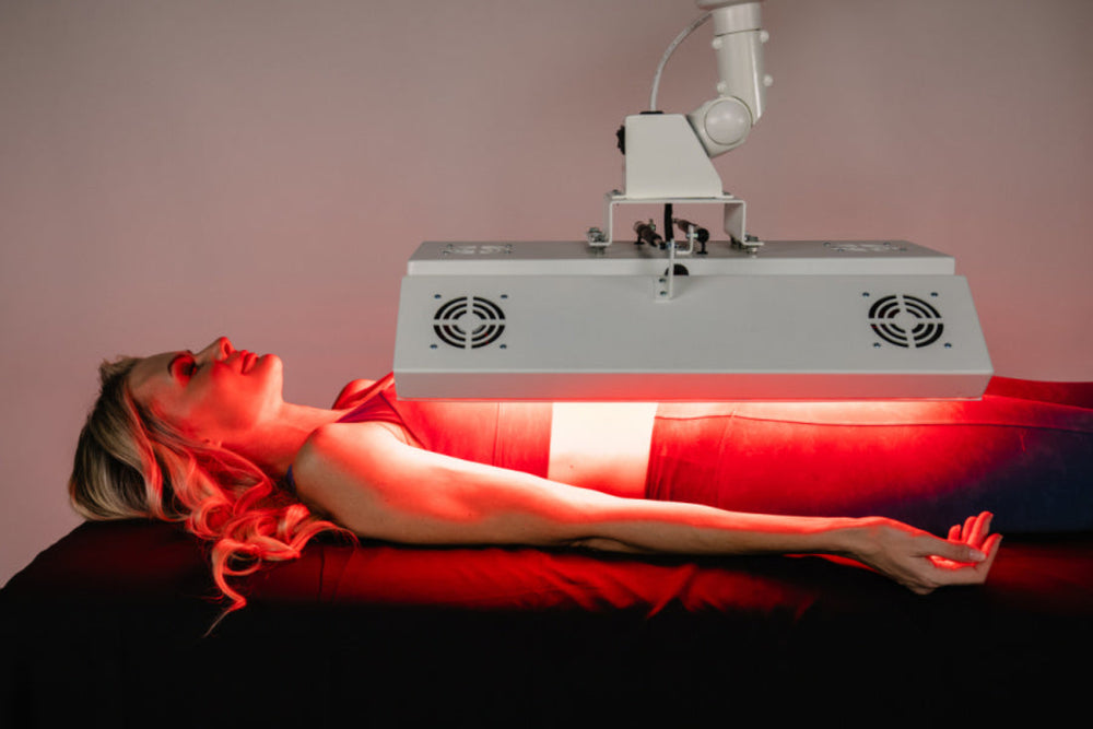 Person receiving red light therapy under a machine with a pink background